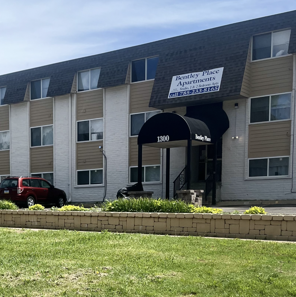Apartment building with a black awning and sign; red car parked in front.