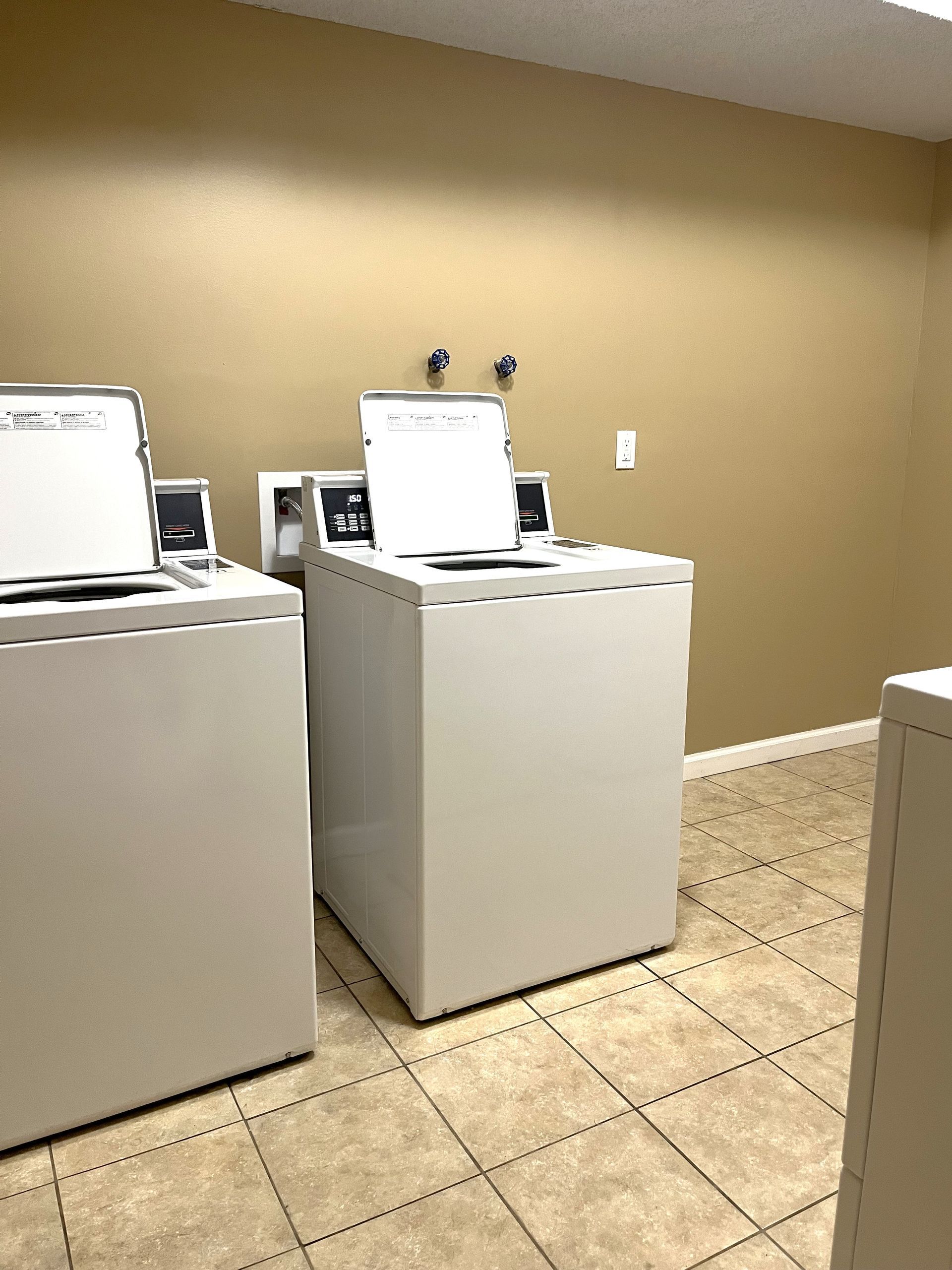 Laundry room with three white washing machines against a beige wall and tiled floor.