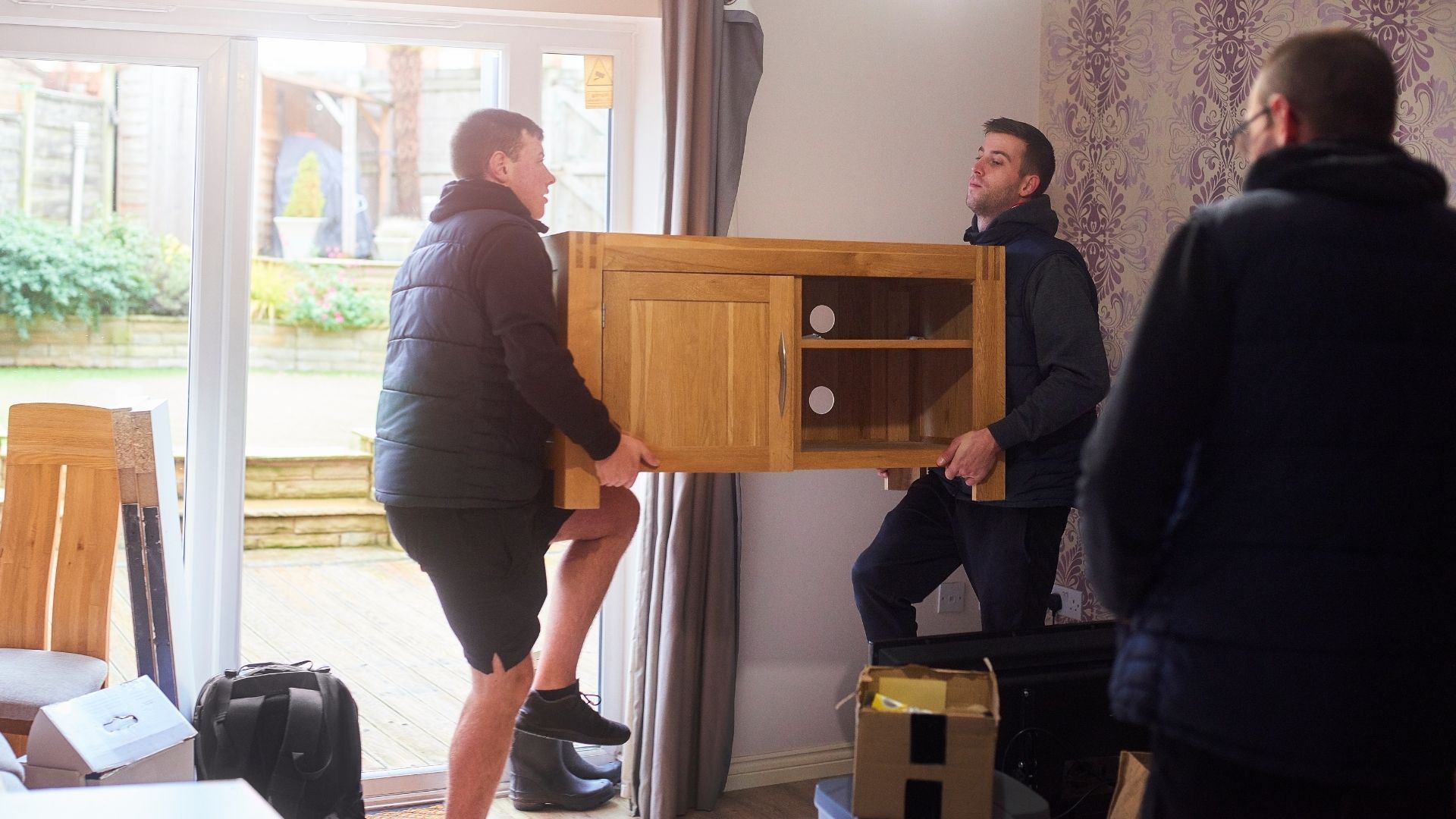 Two men are carrying a wooden cabinet in a living room.