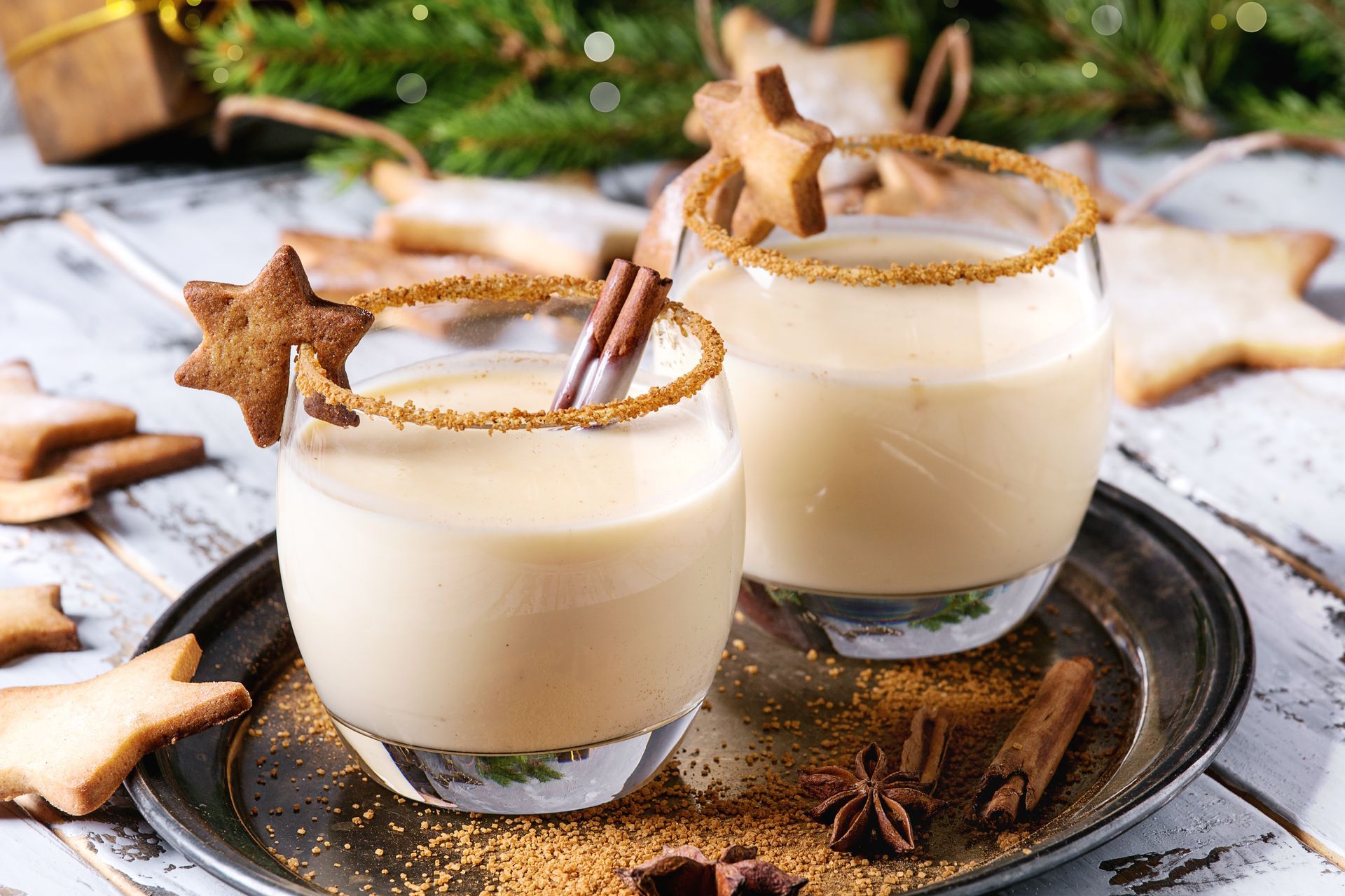 Two glasses of eggnog with star cookies, cinnamon sticks, and sugared rims, on a tray with holiday decorations. Two glasses of eggnog with star cookies, cinnamon sticks, and sugared rims, on a tray with holiday decorations.