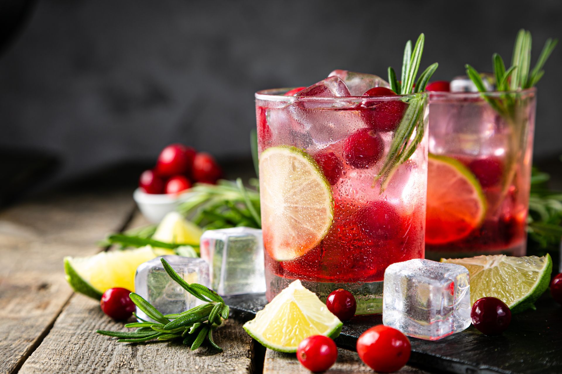 Two cranberry lime cocktails with ice, rosemary sprigs, and lime slices on wooden surface.