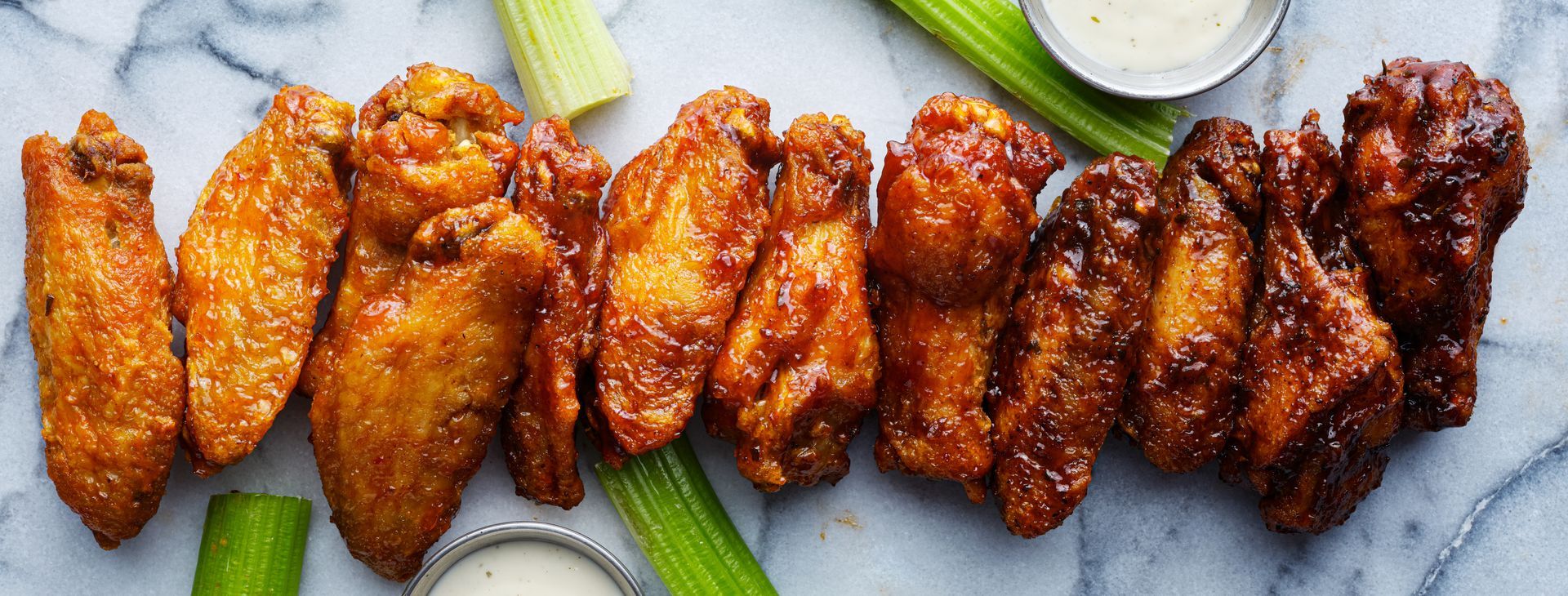 A line of varied, sauced chicken wings arranged on a marble surface with celery sticks and small bowls of dip.