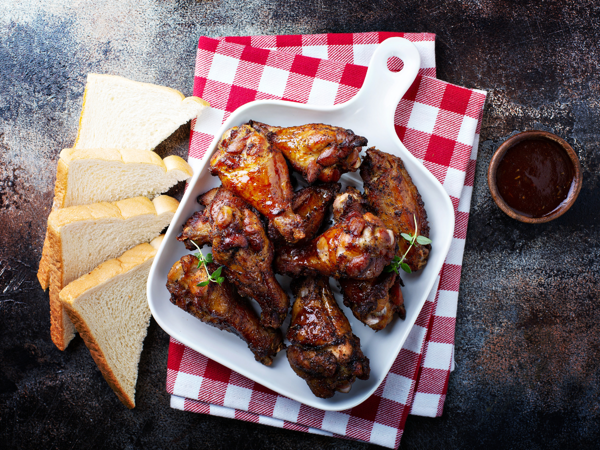 Chicken wings in a white dish on a red and white checkered napkin, with bread slices and a dipping sauce.