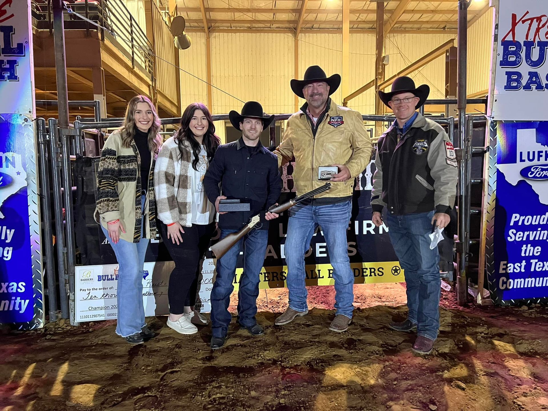 Group of five people posing with a rifle at a rodeo event. They stand inside a dirt arena near a gate.