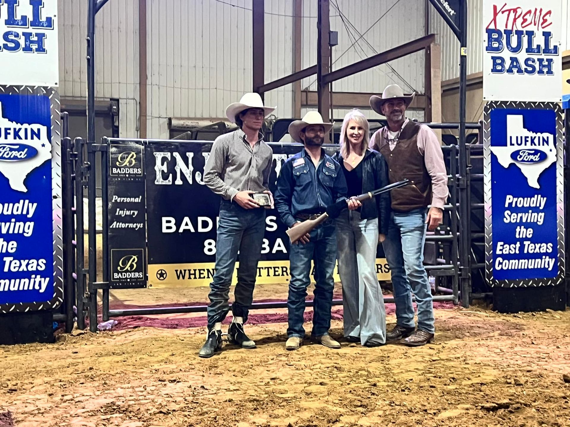 Four people in cowboy attire standing in front of a bull riding arena gate.