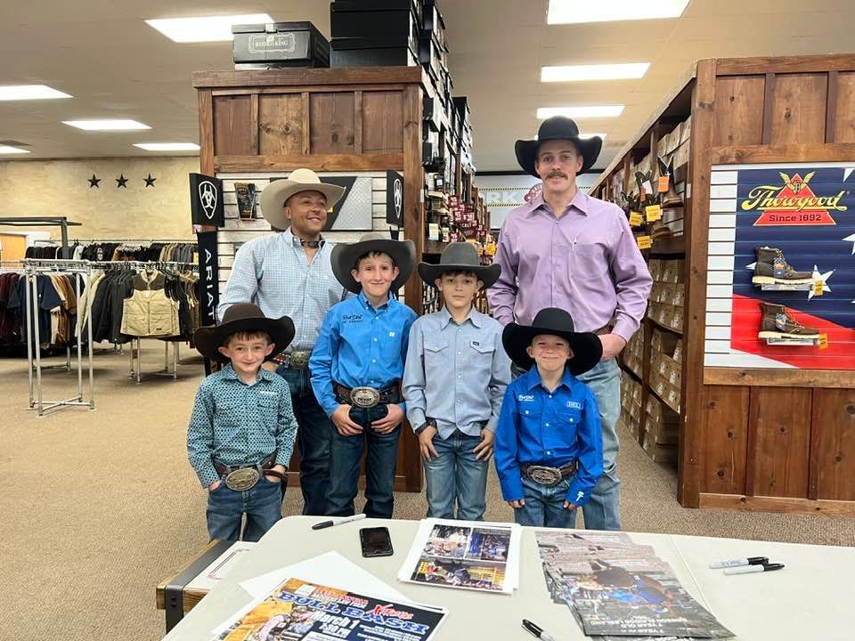 People in cowboy hats and attire pose together. Inside a store, they stand near a table with papers, smiling.
