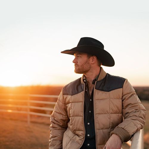 Man in cowboy hat and jacket leans on fence, looking at sunset.