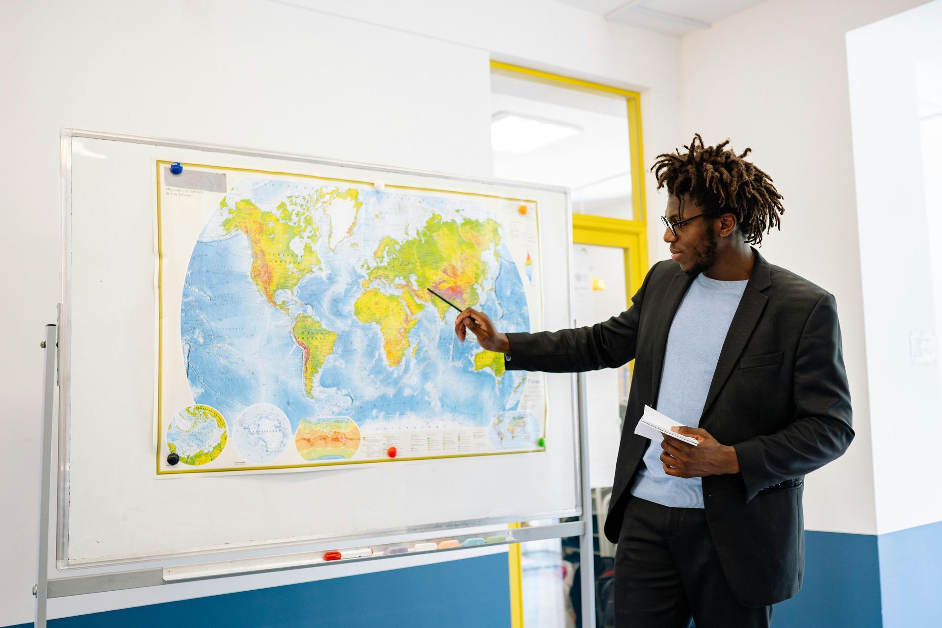 Teacher points to world map on whiteboard. Classroom setting with yellow window.