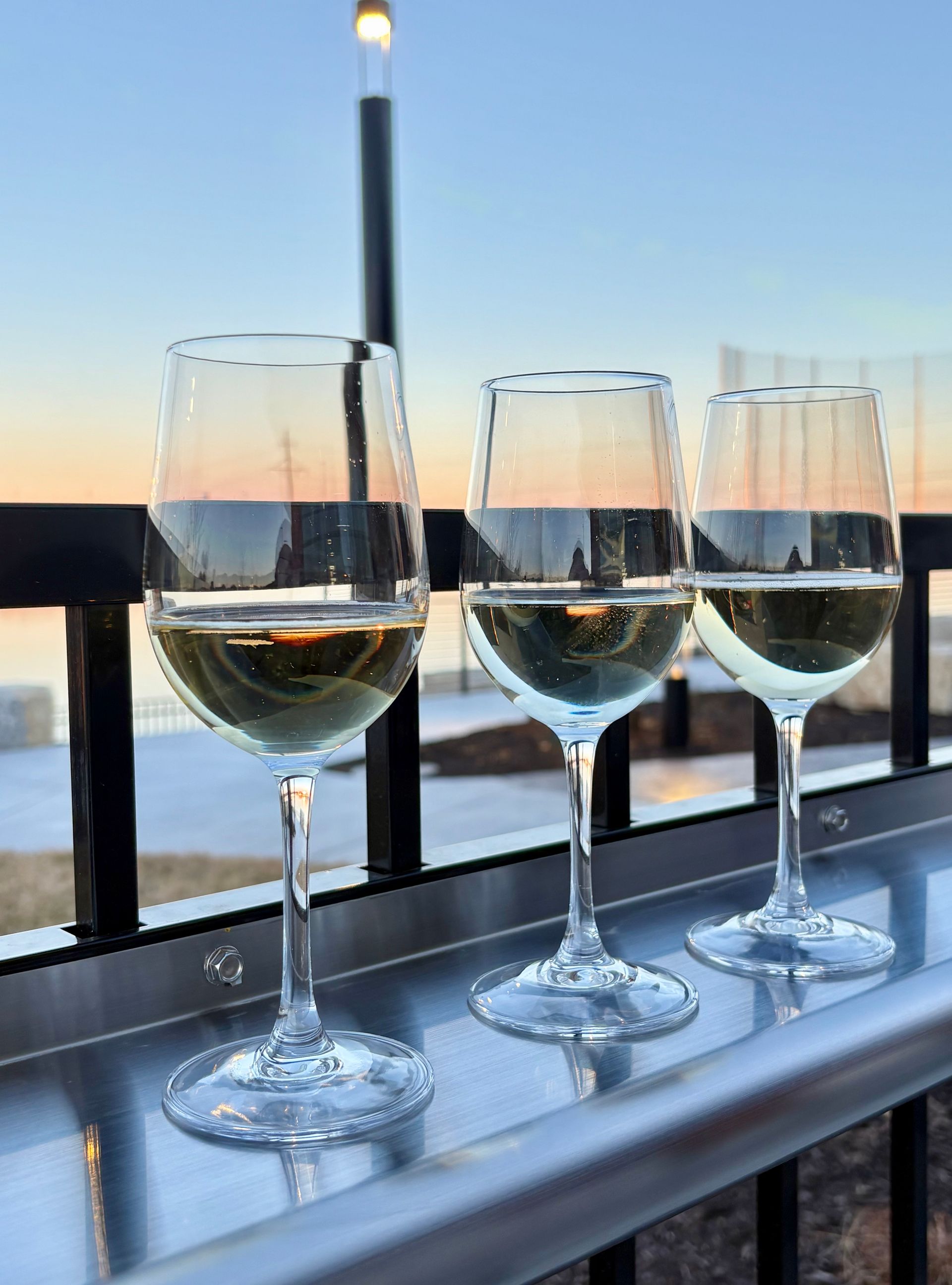 Three glasses of white wine sitting on a metal railing against a scenic sunset background.
