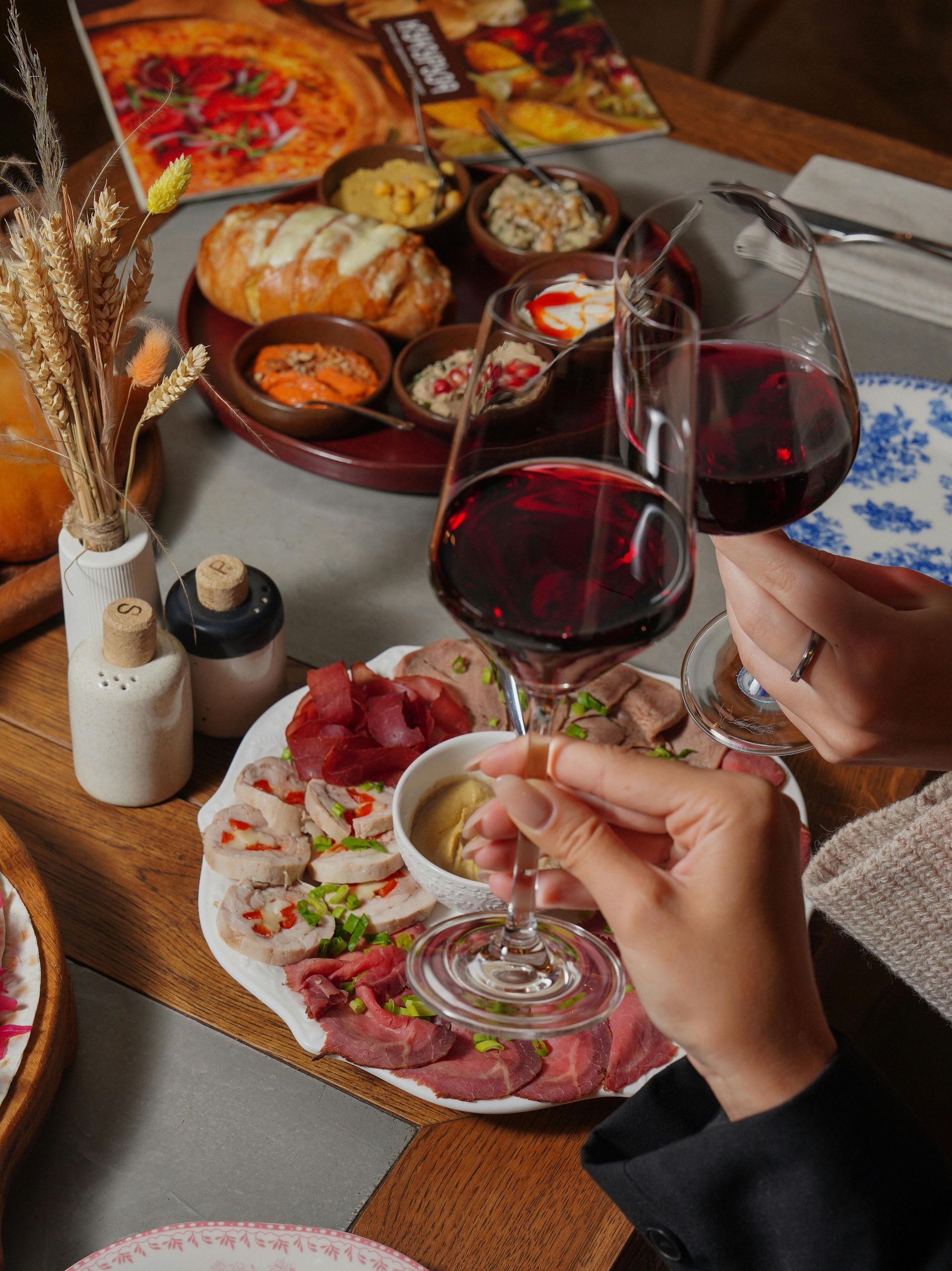 Two wine glasses clink over a charcuterie board and appetizers at a restaurant table.