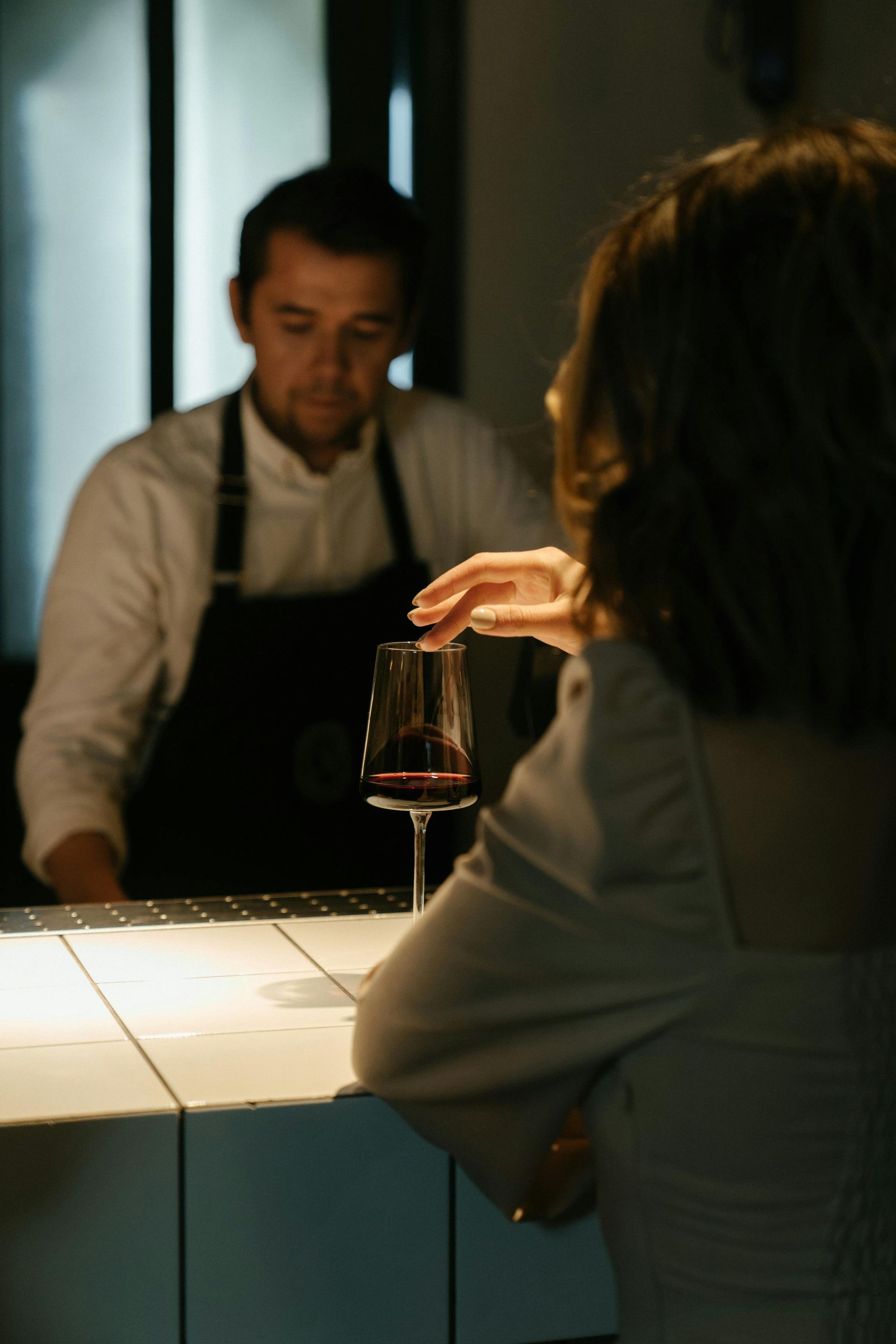 Woman at a bar tasting red wine. Bartender in white shirt and apron watches. Low lighting.