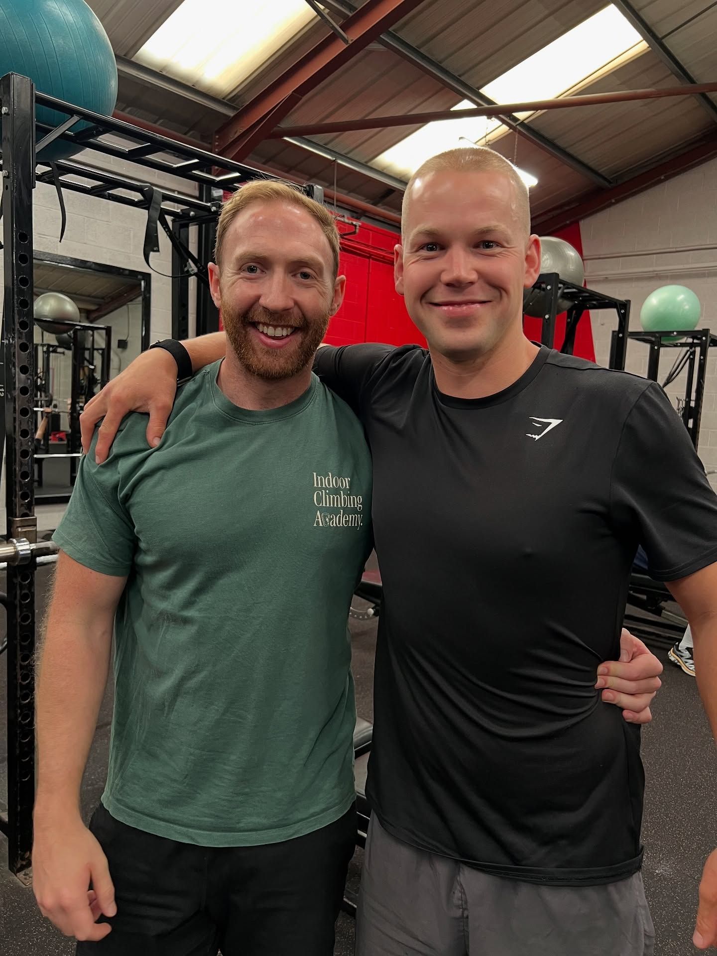 Two smiling people standing arm in arm in a gym in Stockport, with workout equipment in the background.