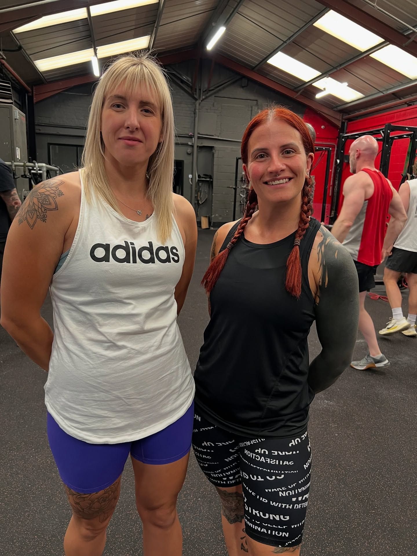 Two women posing in a gym in Stockport, one in white Adidas tank and blue shorts, the other in black top and patterned shorts.