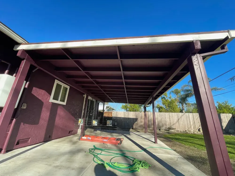 Covered patio with maroon framing, concrete floor, and house on the left. Green hose and orange tool on floor.