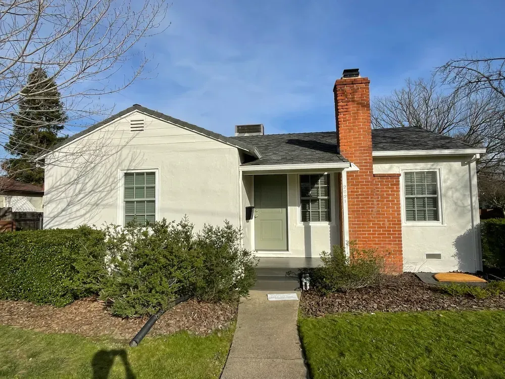 Small white house with brick chimney and front door, green grass, and bushes.