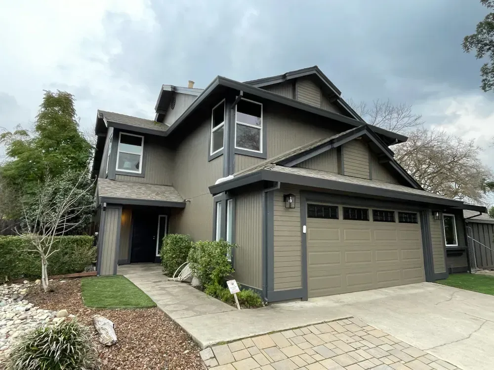 Two-story house, grey exterior, white-framed windows, driveway, and cloudy sky.