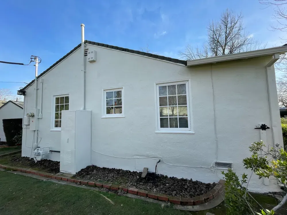 Side view of a light-colored house with white-framed windows, a chimney, and brick edging, under a blue sky.