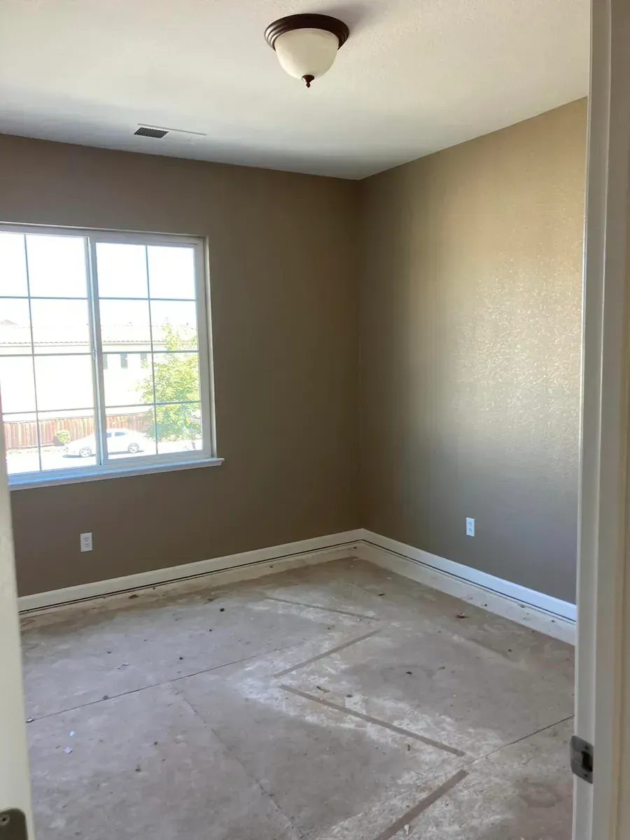 Empty beige room with window and light fixture; dirty floor suggests construction.