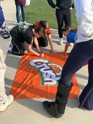 People sign an orange Gatorade towel on pavement; one person wears a medical boot.