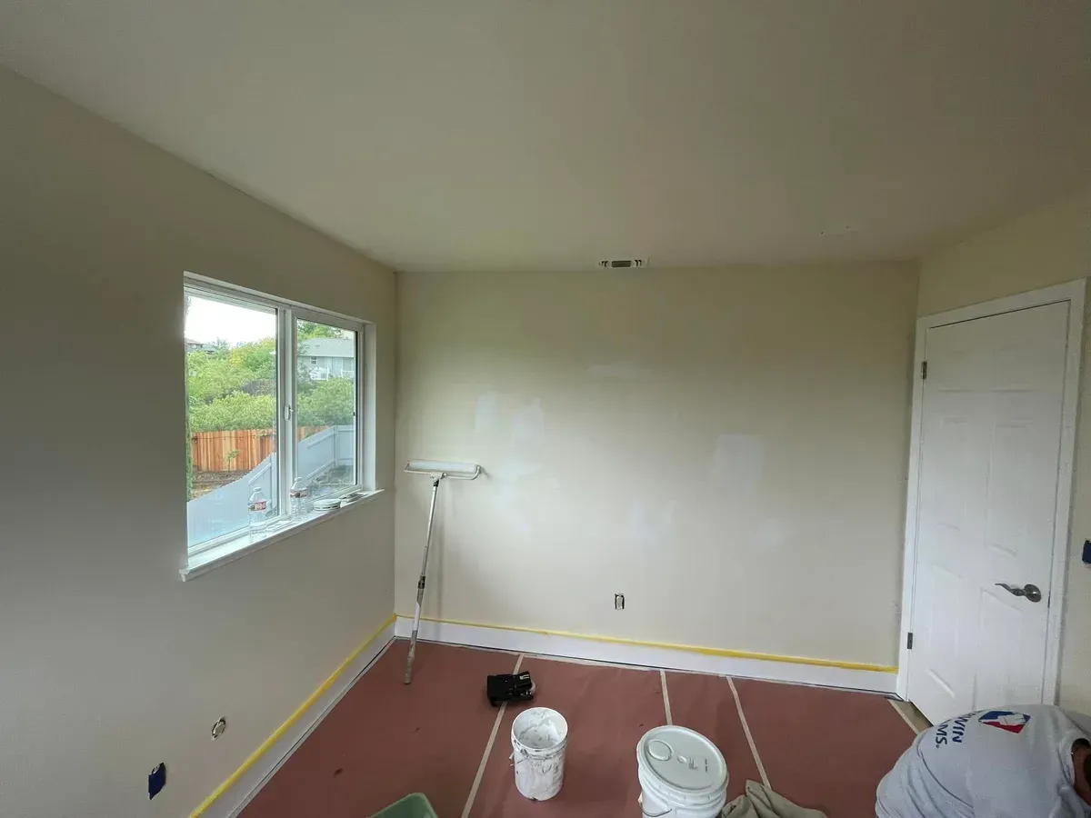 Room with cream-colored walls and a white door/window, preparing for painting, buckets and roller on the floor.
