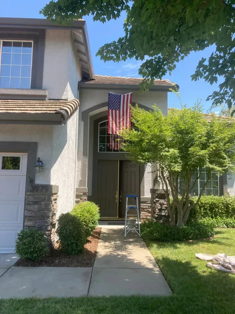 A house with an American flag displayed, a ladder near the front door, and green bushes.