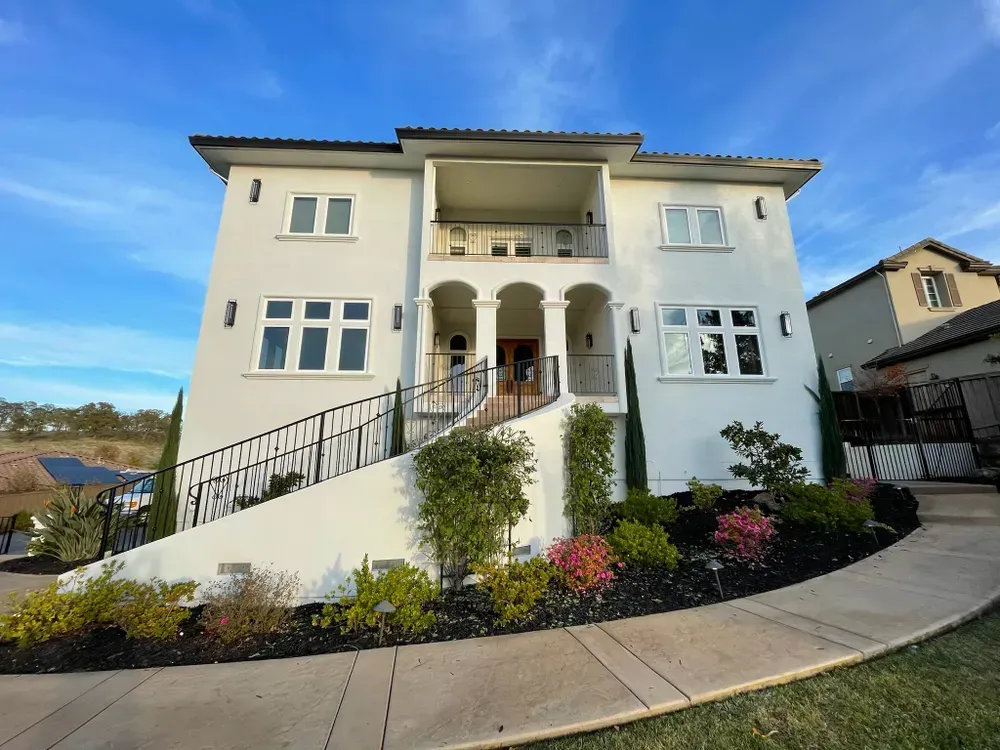 White two-story house with black accents, arched entrance, and a winding pathway. Landscaping with green and red plants.