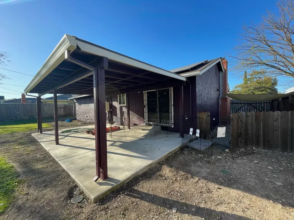 A backyard with a concrete patio, covered by a wooden structure. House with a sliding door and chimney. Sunny day.