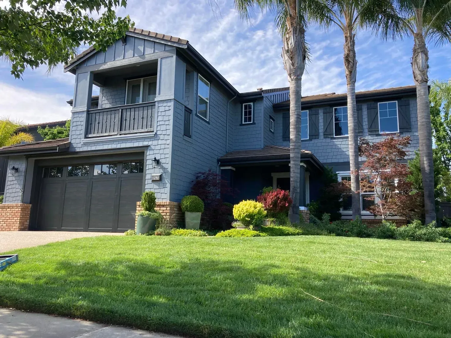 Two-story blue house with green lawn, palm trees, and shrubs. Grey garage door and balcony.