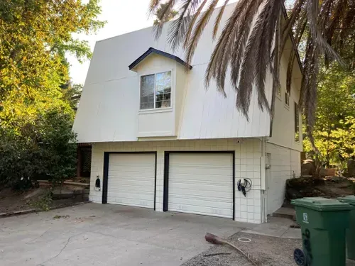Two-story white house with two garage doors. Trees in the background, green trash cans on the right.