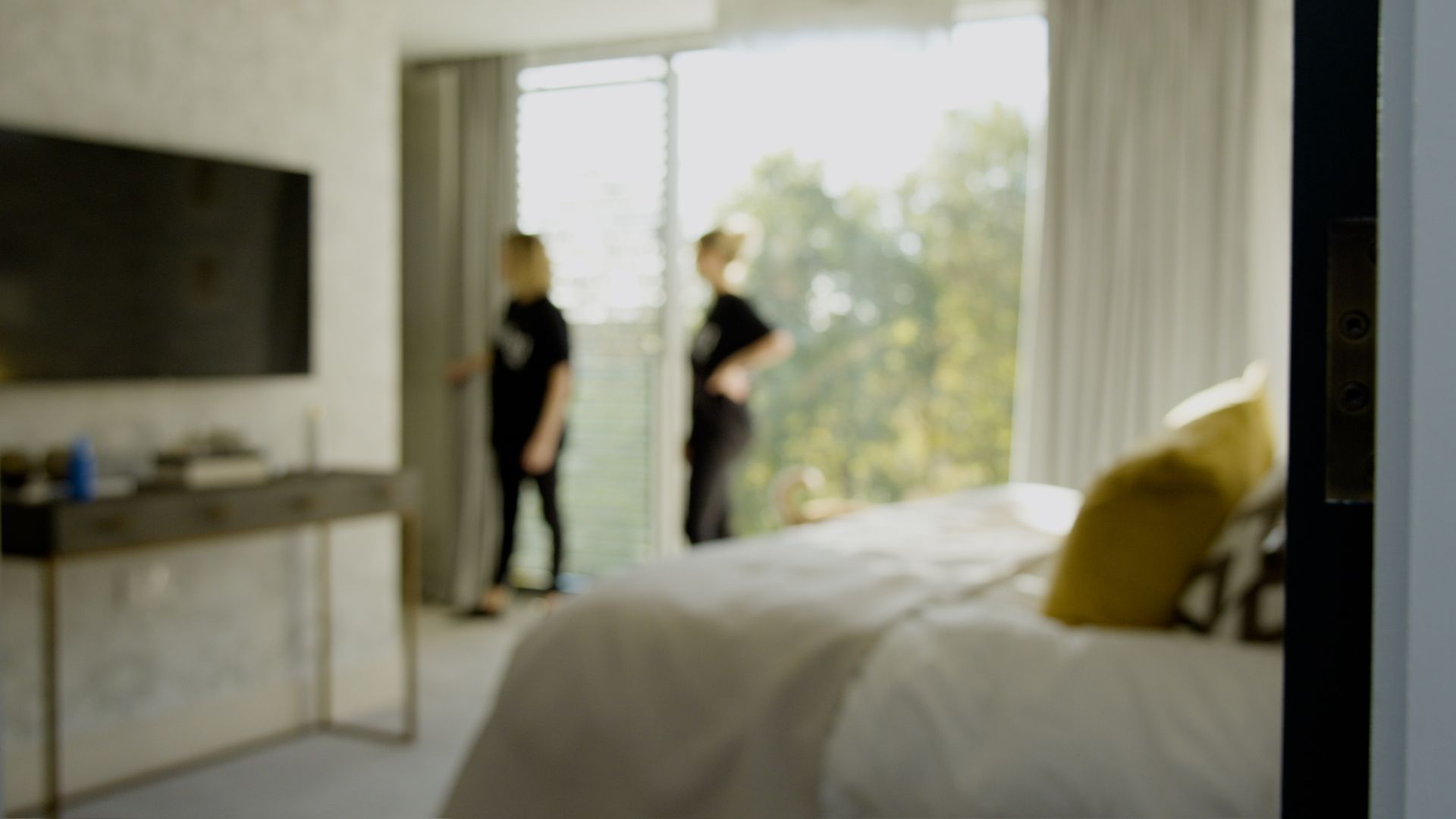 Two people in black clothing stand by a window in a bright bedroom. A bed and TV are visible.