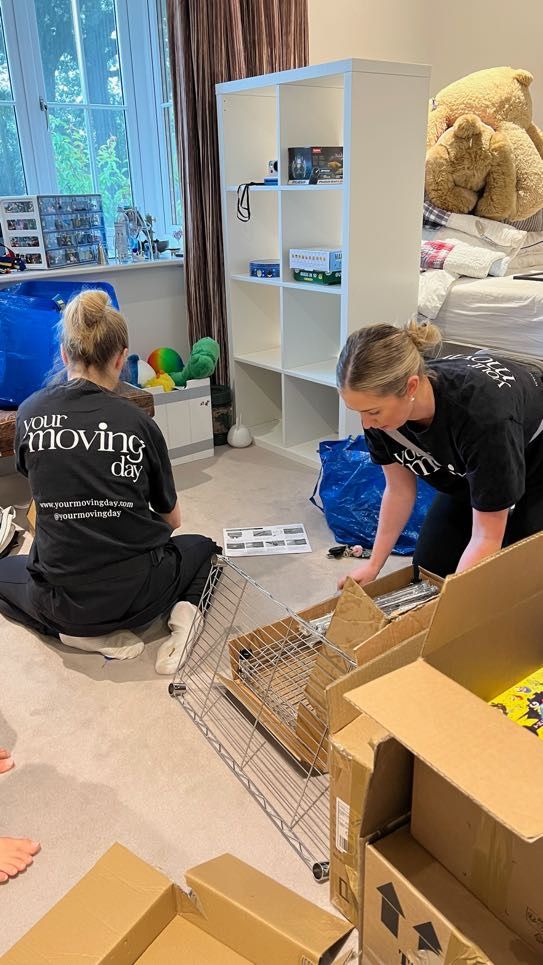 Two people assembling furniture in a room with a window, boxes, and a shelf unit.