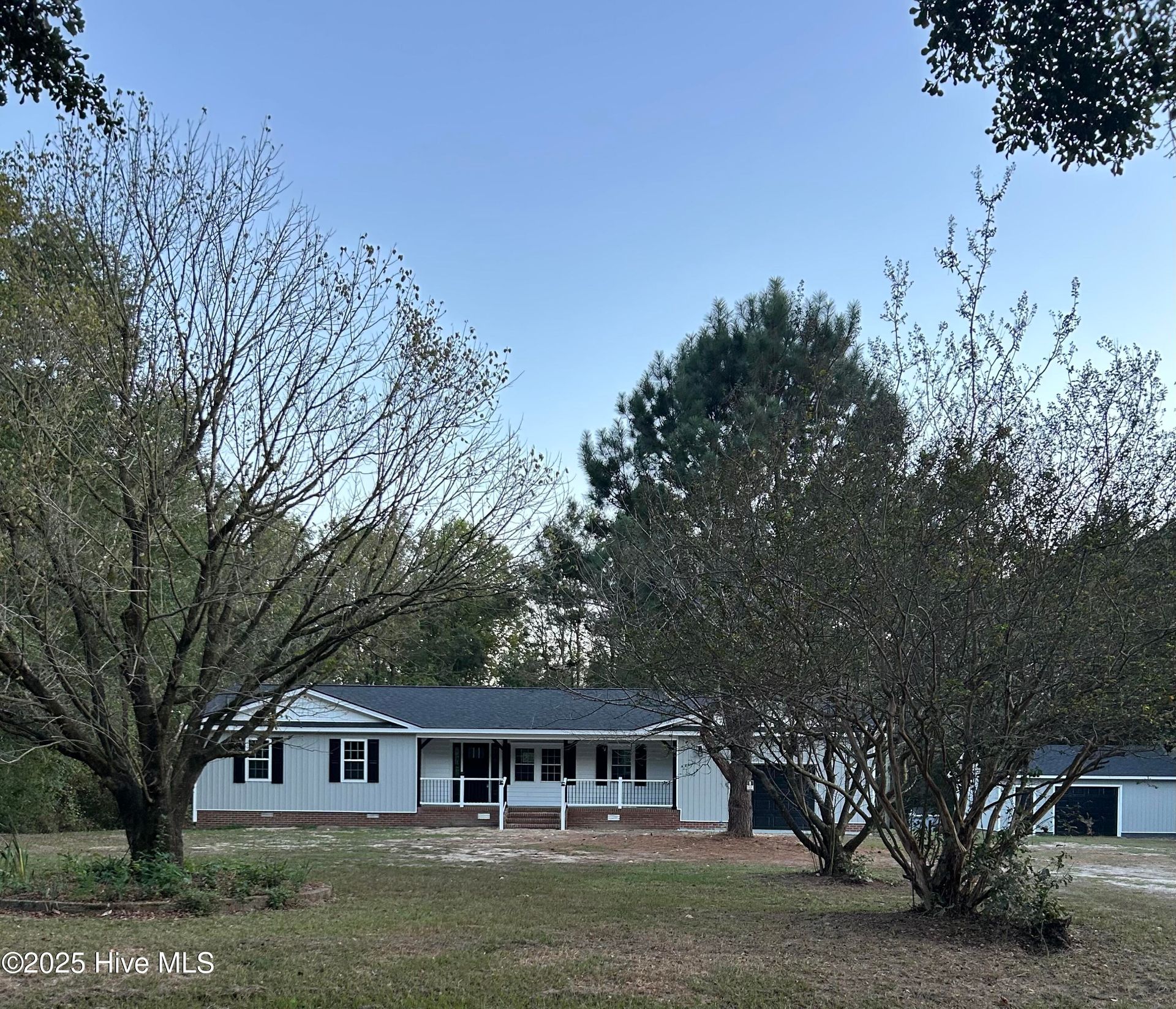 Single-story white house with a porch, partly obscured by trees in a grassy yard under a clear blue sky