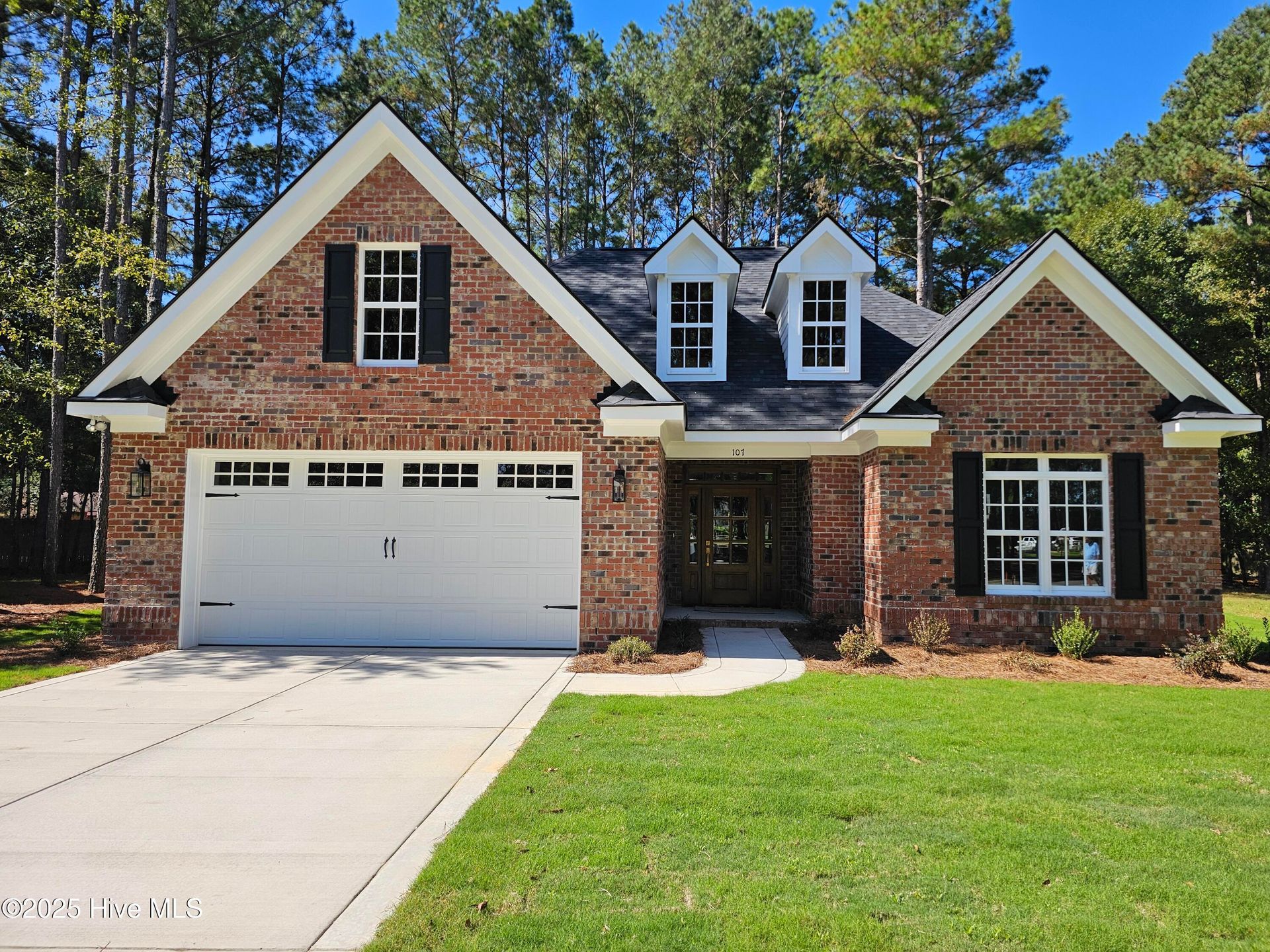 Brick suburban house with white garage, front porch, and dormer windows surrounded by green lawn and trees