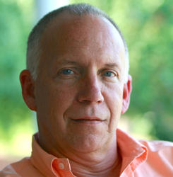Close-up portrait of a man in a peach shirt, sitting outdoors with a blurred green background.