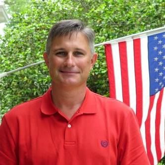 Man in red polo shirt standing outdoors beside a hanging American flag