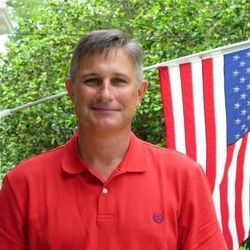 Man in red polo shirt standing outdoors beside a hanging American flag