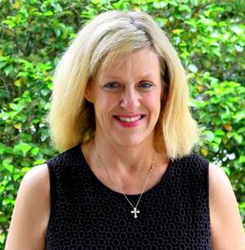Smiling blonde woman in a black sleeveless top, standing outdoors before green foliage