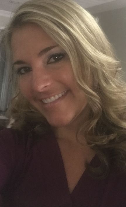 A person with blonde, wavy hair smiles at the camera, wearing a deep purple top in a dimly lit indoor setting.