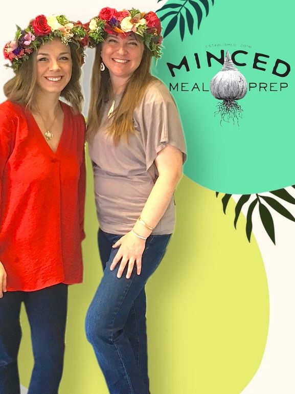 Two women wearing flower crowns are posing for a picture in front of a sign that says mincelia meal prep