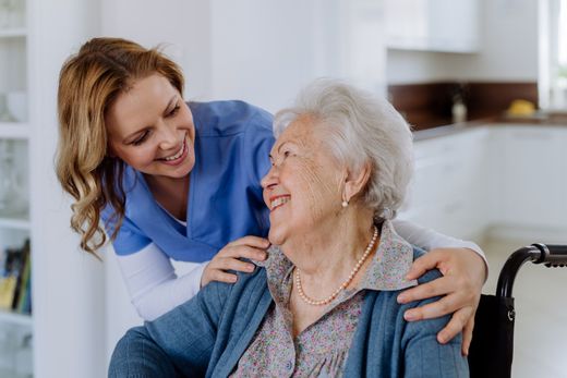 A caregiver in blue scrubs smiles warmly while placing their hands on the shoulders of an older adult in a wheelchair.