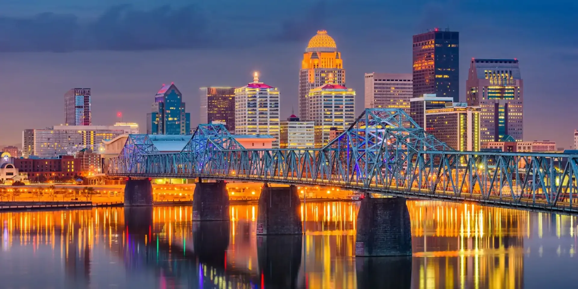 Night view of Louisville, Kentucky skyline with a bridge and water reflection.