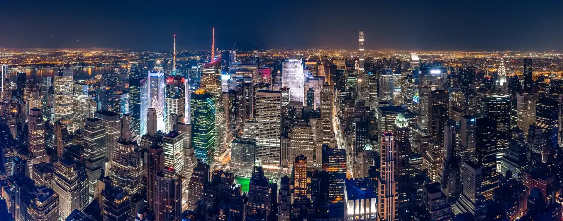 Night view of a cityscape, with bright lights illuminating many tall buildings against a dark blue sky.
