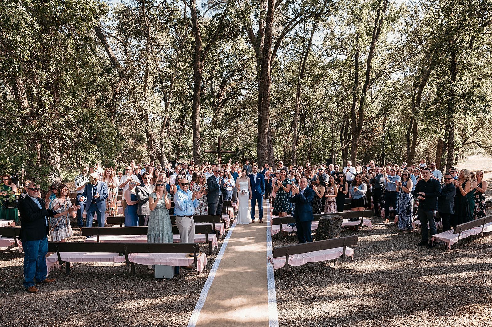 A large group of people are standing in a forest watching a wedding ceremony.