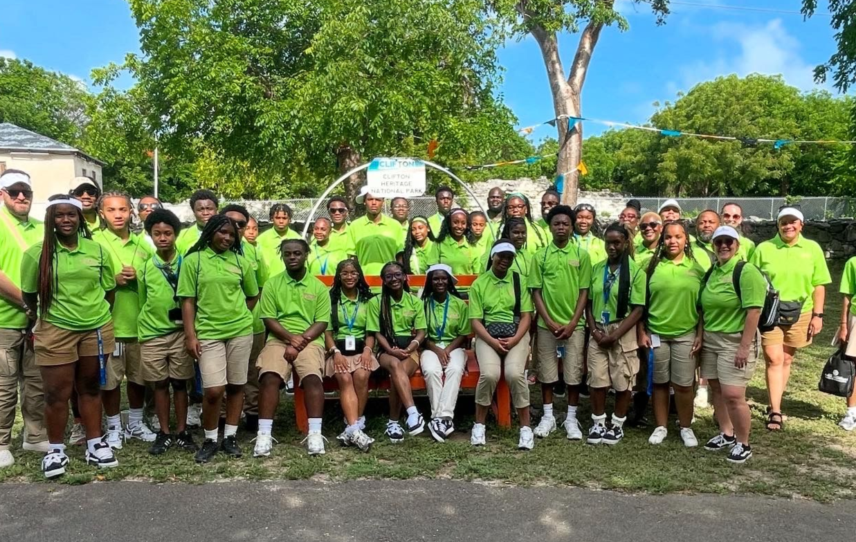 Group of people in green shirts and khaki shorts standing in front of a bench, trees, and a blue sky.