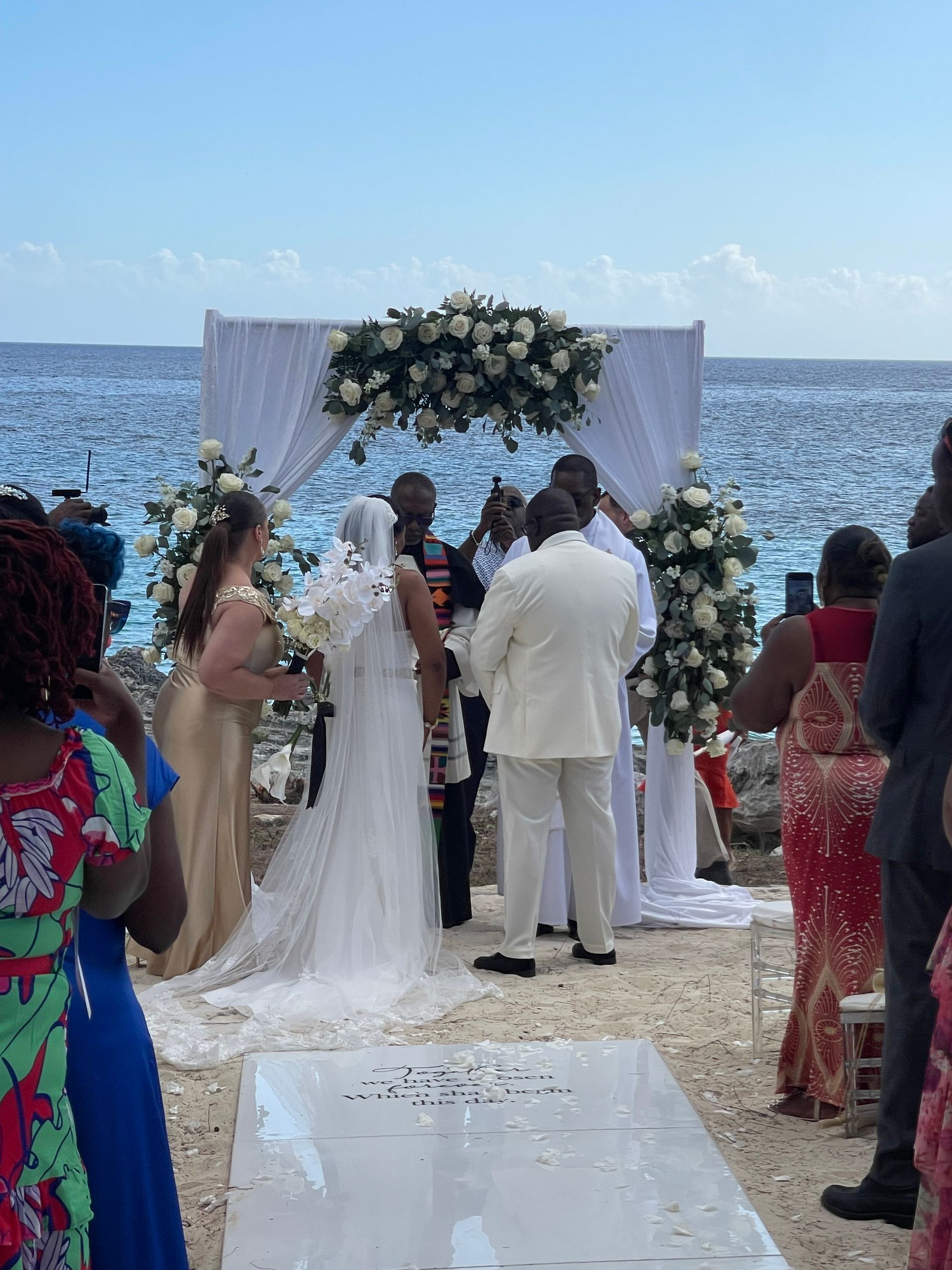 Wedding ceremony on a beach; couple under a floral arch, officiant speaking. Guests watch.