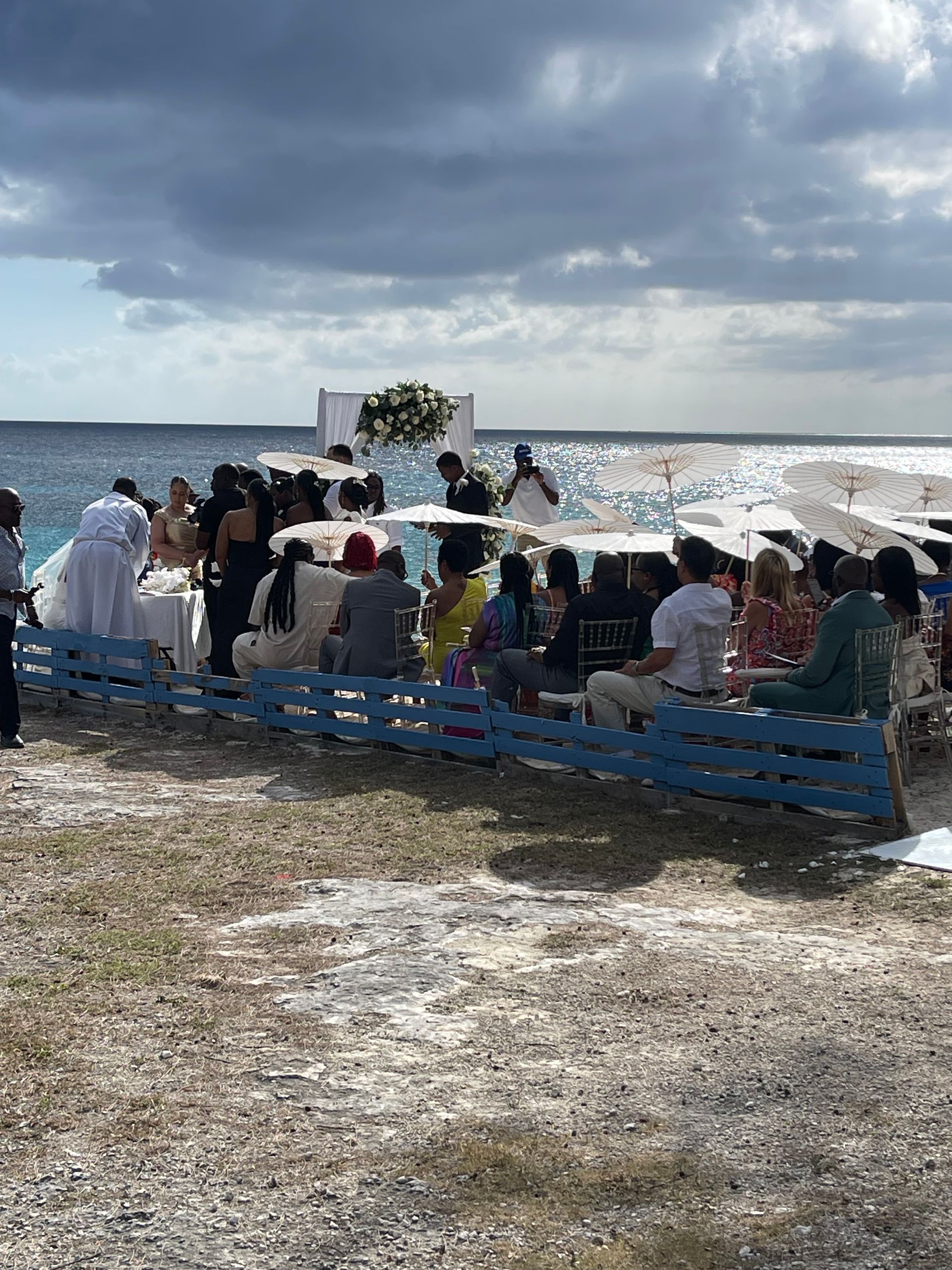 Beach wedding ceremony with guests seated, bride near an arch, ocean in the background.