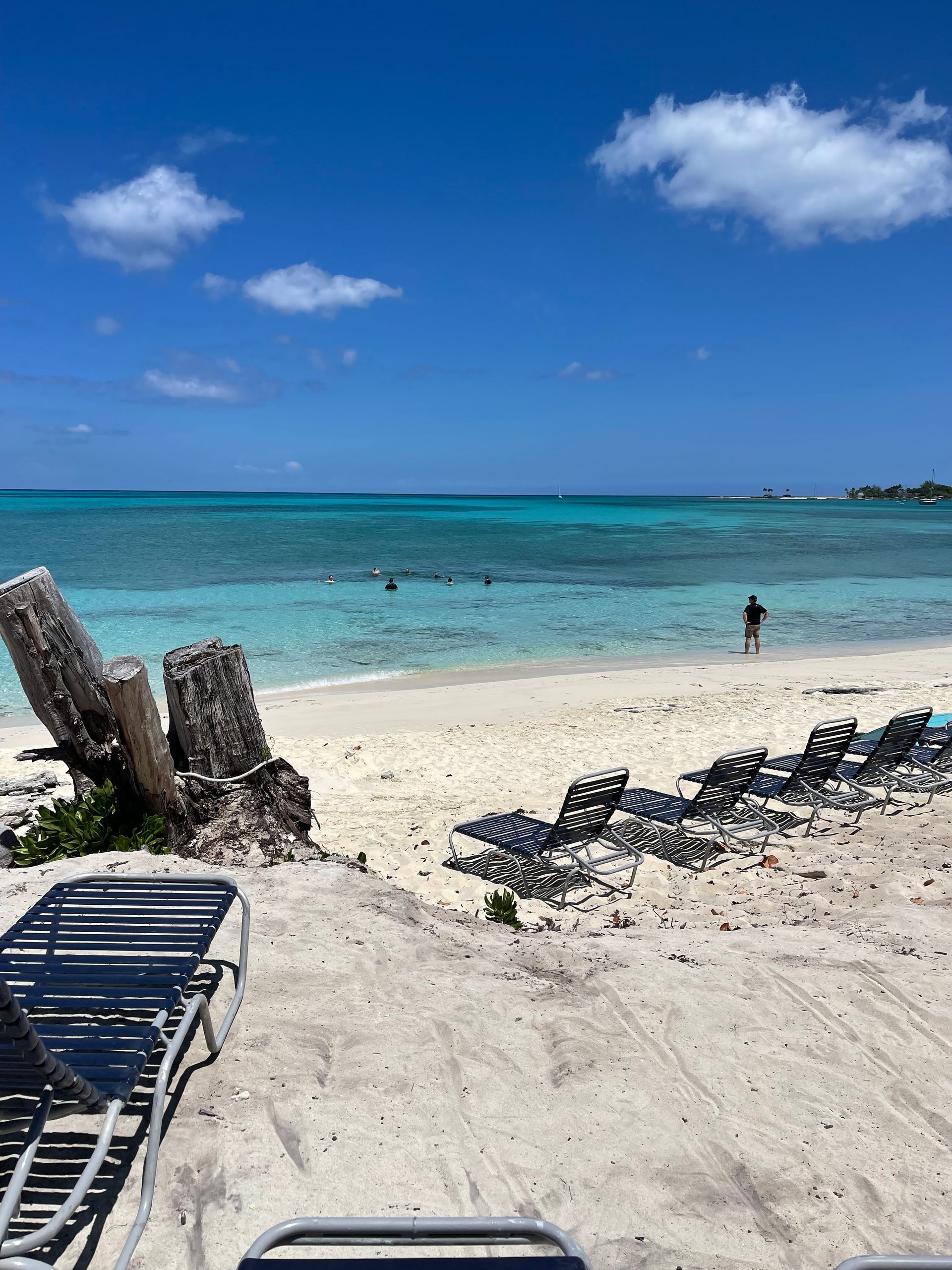 Beach scene with turquoise water, white sand, and blue sky; a few people are in the water.