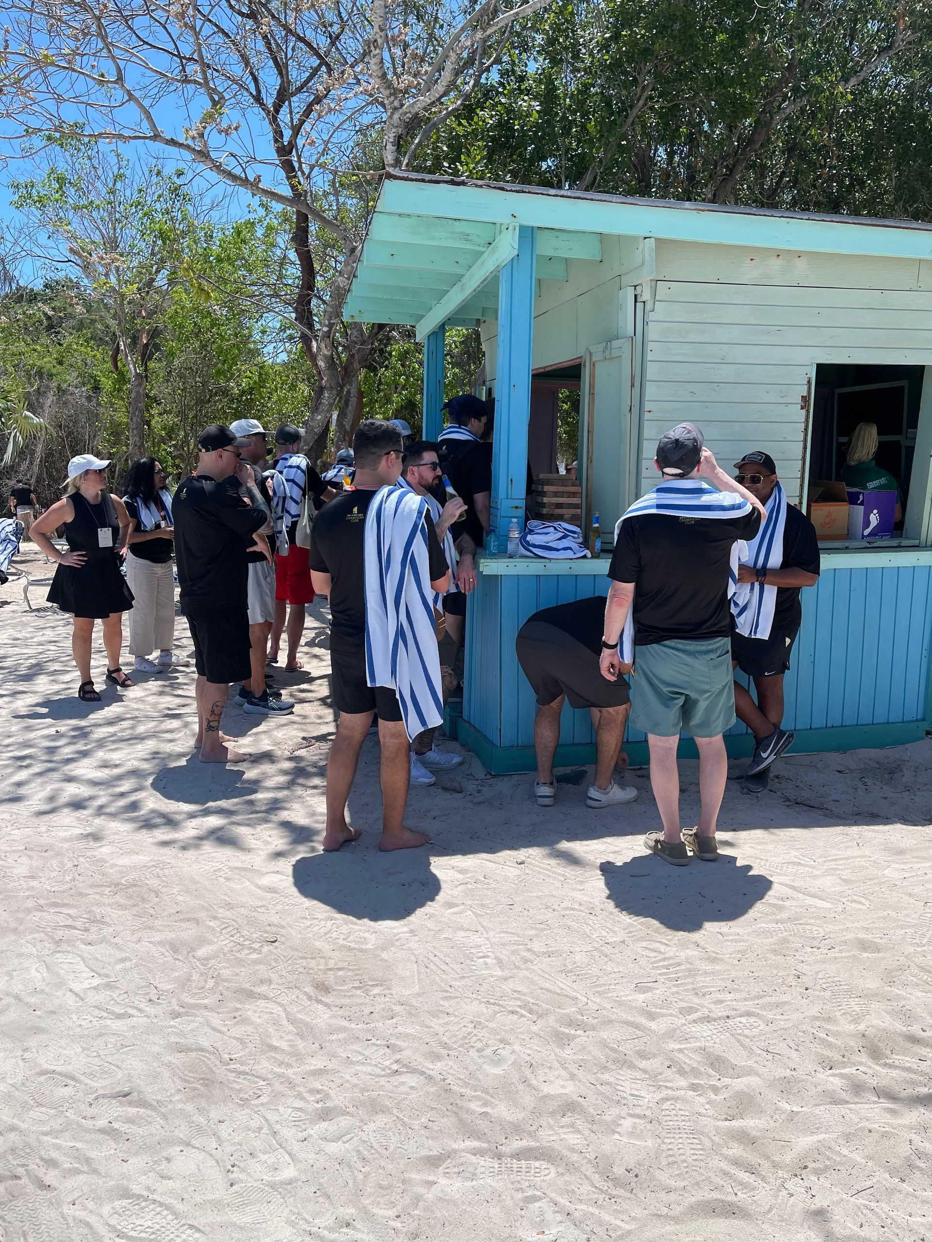People gather around a light blue beach shack on a sunny day. Some are buying items.