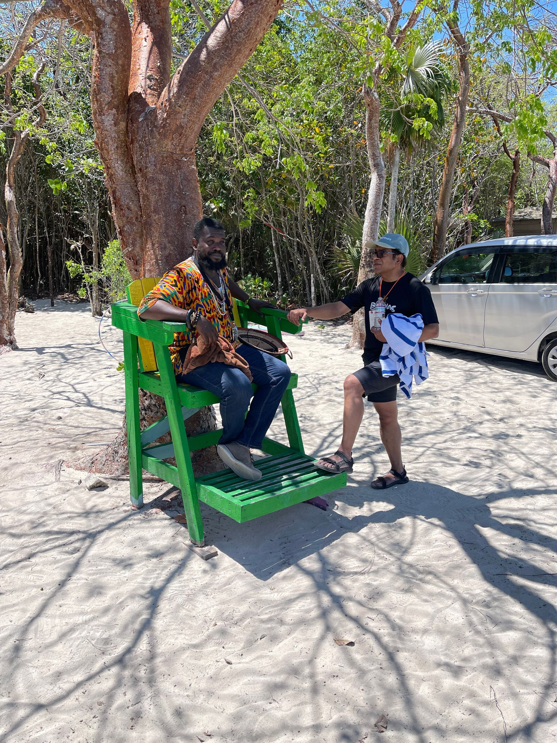 Man seated on green chair; person offering a towel. Beach setting. Sunny day.