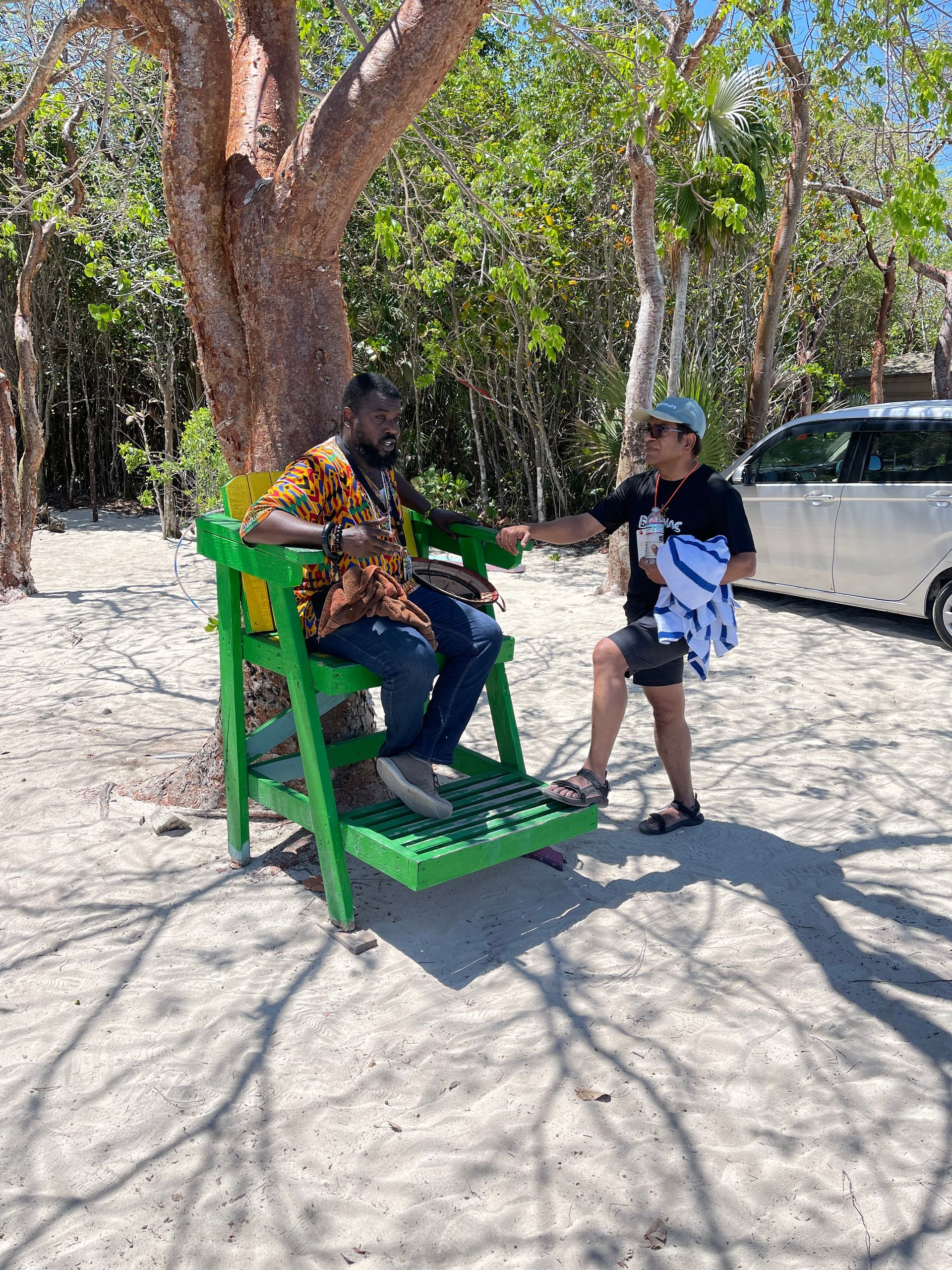 Man seated in green wooden chair shakes hands with another person near a car under trees.